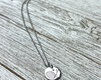 Puede incluir: Un collar de plata con un colgante redondo que presenta un diseño dibujado a mano de una bola de hilo y esquís. El colgante está unido a una cadena de plata.