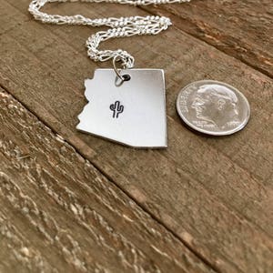 May include: A silver necklace with a pendant shaped like the state of Arizona. The pendant features a small cactus design. The necklace is on a wooden surface next to a US dime.