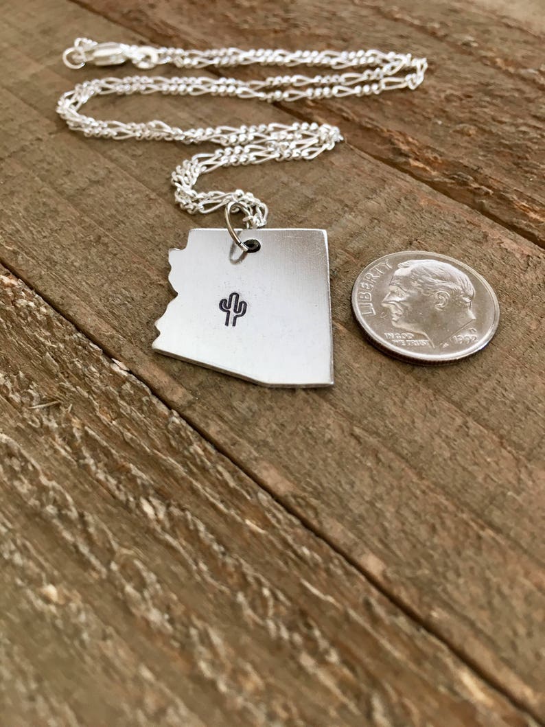 May include: A silver necklace with a pendant shaped like the state of Arizona. The pendant features a small cactus design. The necklace is on a wooden surface next to a US dime.