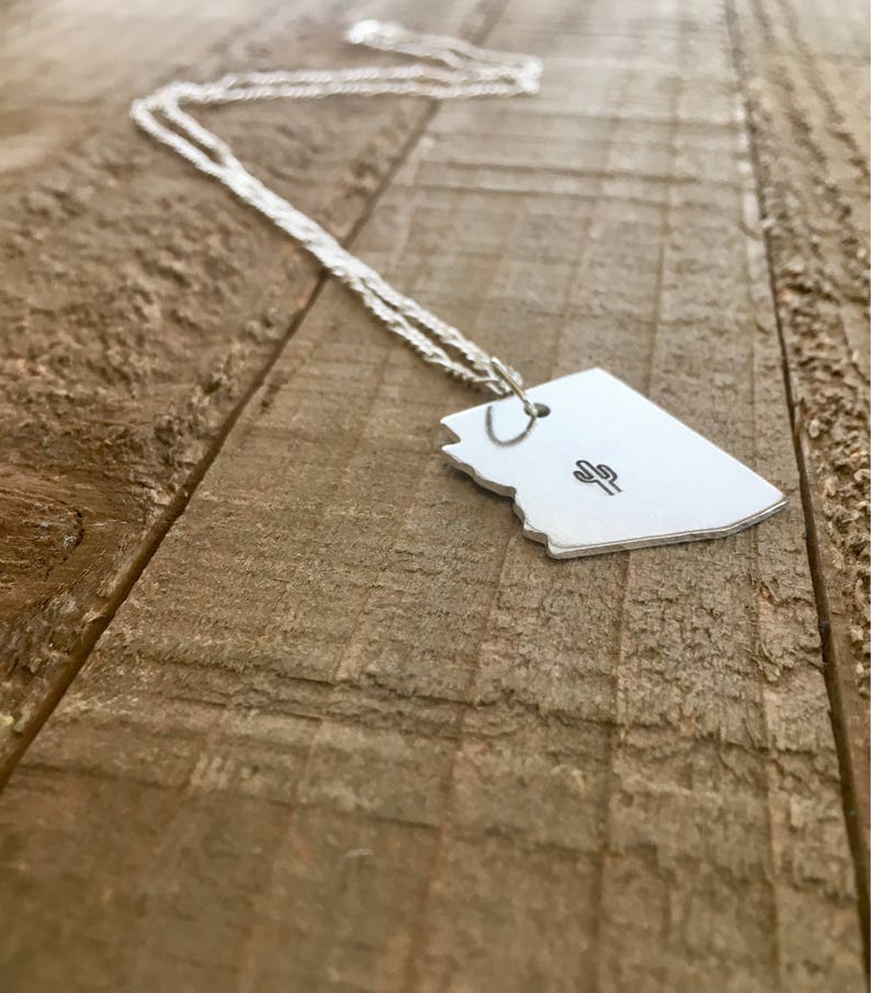 May include: A silver necklace with a pendant shaped like the state of Arizona featuring a small cactus design.