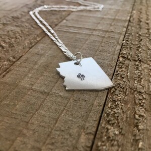 May include: A silver necklace with a pendant shaped like the state of Arizona featuring a small cactus design. The necklace is displayed on a wooden surface.