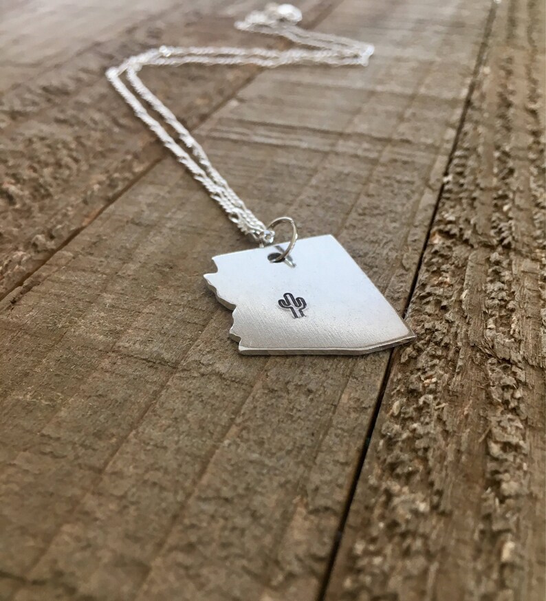 May include: A silver necklace with a pendant shaped like the state of Arizona featuring a small cactus design. The necklace is displayed on a wooden surface.