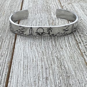 Op de afbeelding: Een zilveren metalen armband met een gegraveerd ontwerp van een palmboom, zonsondergang, golven en surfplanken.