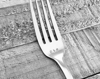 May include: A silver fork with the word "pie" engraved on the handle. The fork is lying on a wooden surface.