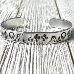 May include: A silver metal bracelet with a design of cacti, aliens, and a mountain range. The bracelet is on a wooden surface.