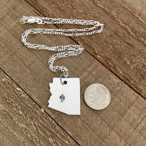 May include: A silver necklace with a pendant shaped like the state of Arizona featuring a small cactus design. The pendant is approximately the size of a 10 pence coin.