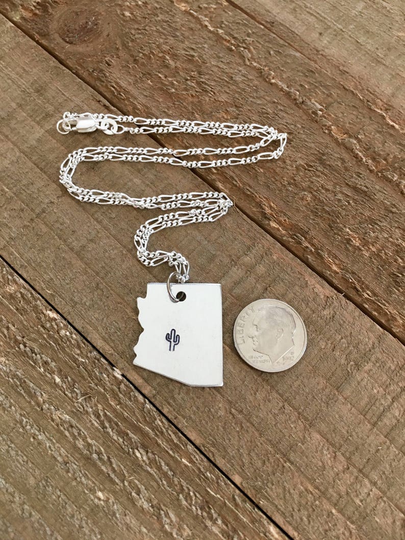 May include: A silver necklace with a pendant shaped like the state of Arizona featuring a small cactus design. The pendant is approximately the size of a 10 pence coin.