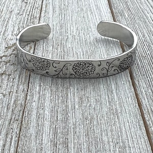 May include: A silver bangle bracelet with an engraved floral design. The design features delicate vines and stylized flowers.