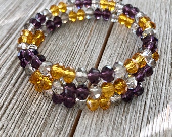 Puede incluir: Un brazalete de triple hebra hecho con cuentas de vidrio transparentes, moradas y doradas. El brazalete está envuelto alrededor de un alambre de plata.