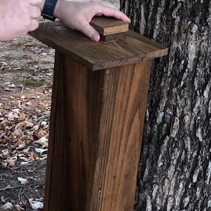 May include: A wooden bird feeder with a rectangular design and a small roof. The feeder is made of stained wood and is mounted on a tree trunk. The feeder has a compartment at the bottom filled with birdseed.