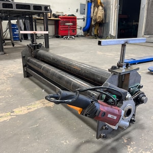 May include: A metal pipe bending machine with a red and black electric motor attached to the side. The machine has three rollers and a hand crank.