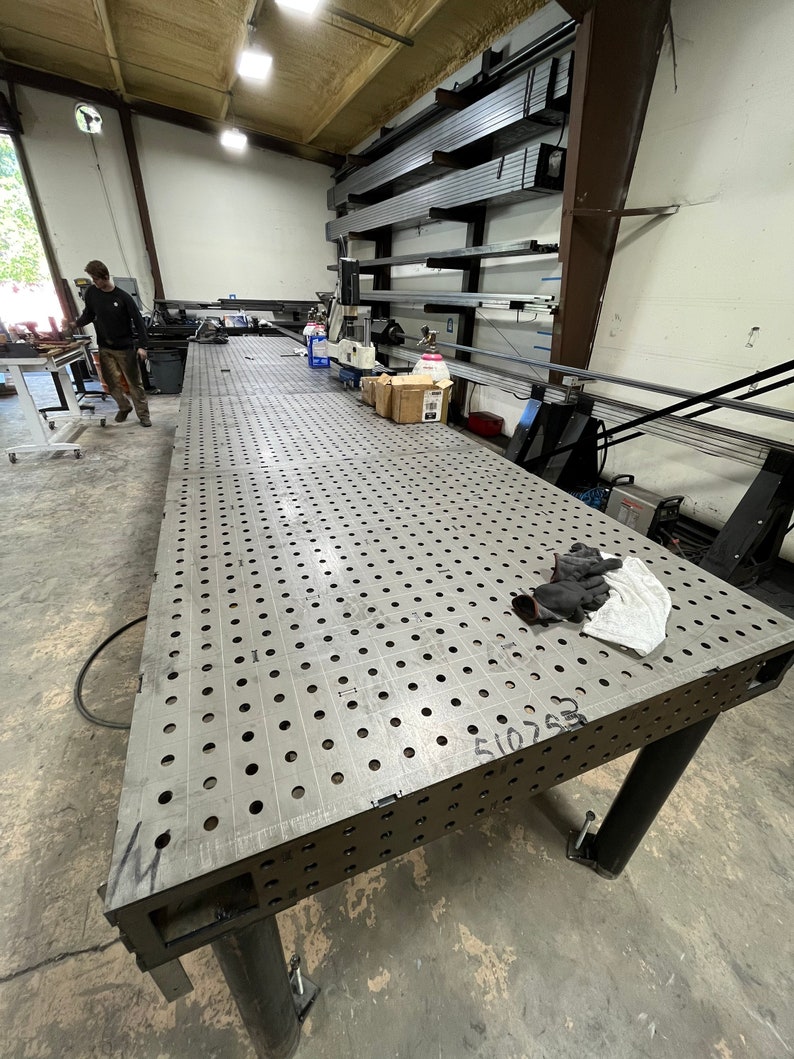 May include: A large metal welding table with a perforated surface and a black number "510763" written on the surface. The table has four black legs and is in a workshop setting.