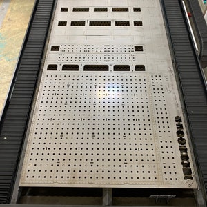 May include: A metal sheet with a grid of holes and several rectangular cutouts. The sheet is on a black platform with a blue and black machine in the background. The text "551" is written on the sheet.