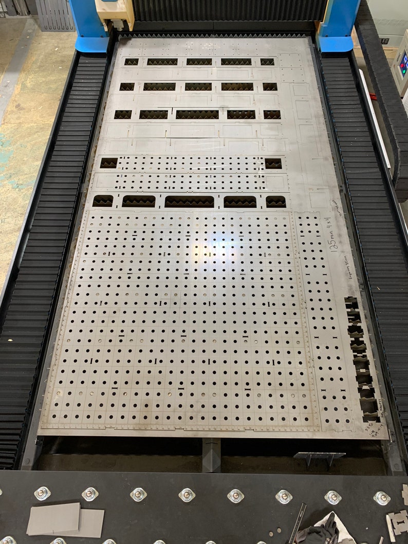 May include: A metal sheet with a grid of holes and several rectangular cutouts. The sheet is on a black platform with a blue and black machine in the background. The text "551" is written on the sheet.