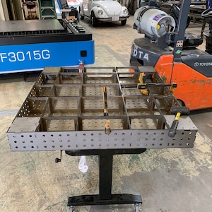 May include: A metal welding table with a grid of squares and holes. The table is on a black adjustable stand with wheels. The table is in a workshop setting with a forklift and a laser cutter in the background.