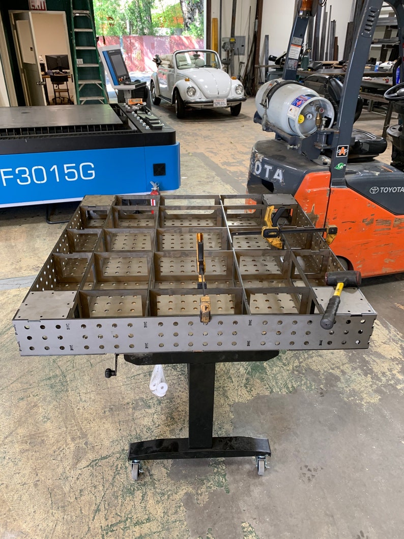 May include: A metal welding table with a grid of squares and holes. The table is on a black adjustable stand with wheels. The table is in a workshop setting with a forklift and a laser cutter in the background.