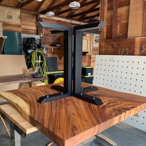 May include: A large, live edge wooden table top with a natural wood grain pattern. The table top is supported by a black metal base with a unique, geometric design.