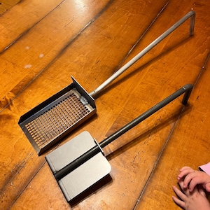 May include: Two metal fireplace tools with long handles. The tool on the left has a rectangular metal scoop with a wire mesh bottom. The tool on the right has a solid metal scoop.