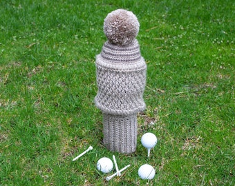May include: A beige knitted golf hat with a brown pom pom on top, sitting on green grass with four white golf balls and tees.