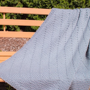 May include: A gray knitted throw blanket with a textured pattern, draped over a wooden bench.