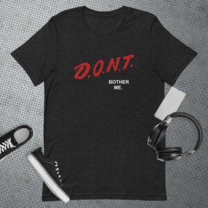 Puede incluir: Camiseta gris oscuro con el texto rojo "D.O.N.T. BOTHER ME." Zapatillas deportivas blancas y negras y auriculares negros también están en la imagen. Un smartphone blanco también está en la imagen.