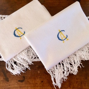 Puede incluir: Dos toallas de mano blancas con bordes con flecos e iniciales bordadas en azul y amarillo "C A".