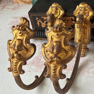 May include: A pair of gold-toned metal curtain tiebacks featuring ornate, symmetrical scrollwork and leaf-like detailing. Each tieback has a curved hook extending from a decorative wall-mounted plate with a distressed, aged finish.