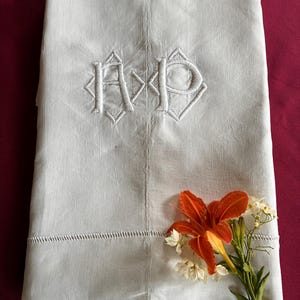 Puede incluir: Un textil blanco doblado que presenta un monograma bordado con las letras AP en un marco geométrico en forma de diamante. La tela tiene un detalle de dobladillo calado cerca del borde inferior, acentuado por un lirio naranja y pequeñas flores blancas.