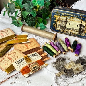 Puede incluir: Una colección de hilos de coser antiguos de varios colores, etiquetas vintage y una caja de metal azul decorativa con la inscripción "Bergamotes de Nancy". Los artículos están dispuestos sobre un paño blanco con bordes de encaje.