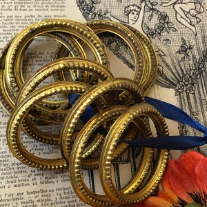 May include: Several gold-colored oval frames with a textured beaded design are grouped together, some with blue ribbon. The frames are arranged on a vintage paper background with floral and botanical illustrations.