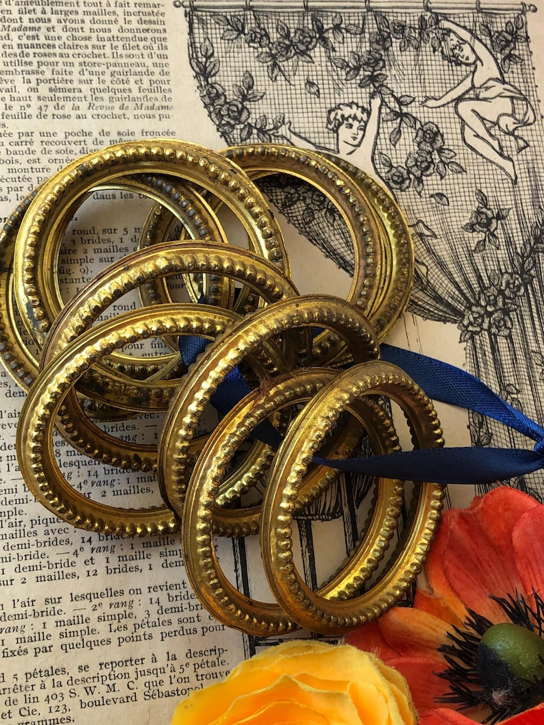 May include: Several gold-colored oval frames with a textured beaded design are grouped together, some with blue ribbon. The frames are arranged on a vintage paper background with floral and botanical illustrations.