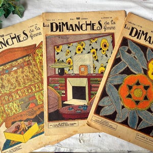 May include: Three vintage French magazines titled "Les Dimanches de la femme" are displayed. The covers feature colorful illustrations, including floral designs and interior scenes. The magazines are aged with visible wear.