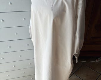 Puede incluir: Un camisón largo blanco con cuello cuadrado y mangas largas. El camisón presenta delicados detalles bordados en el corpiño. La prenda se exhibe en un maniquí, mostrando su diseño fluido y estilo vintage.
