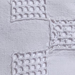 May include: Close-up of white fabric featuring a repeating square openwork embroidery pattern. The embroidery is a delicate, raised design, creating a textured effect against the smooth background. The overall aesthetic is clean and classic.