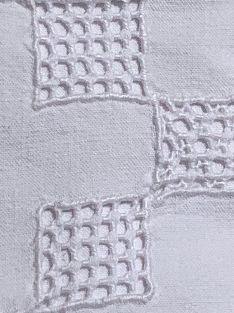May include: Close-up of white fabric featuring a repeating square openwork embroidery pattern. The embroidery is a delicate, raised design, creating a textured effect against the smooth background. The overall aesthetic is clean and classic.