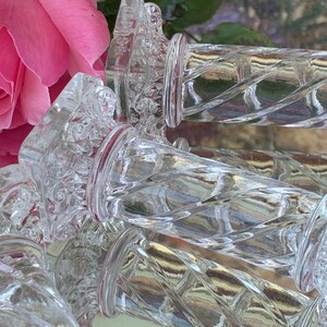 May include: Clear glass bottle stoppers with a decorative, twisted design. The stoppers have a clear, textured body and a red band near the top. A pink rose and green leaves are in the background.