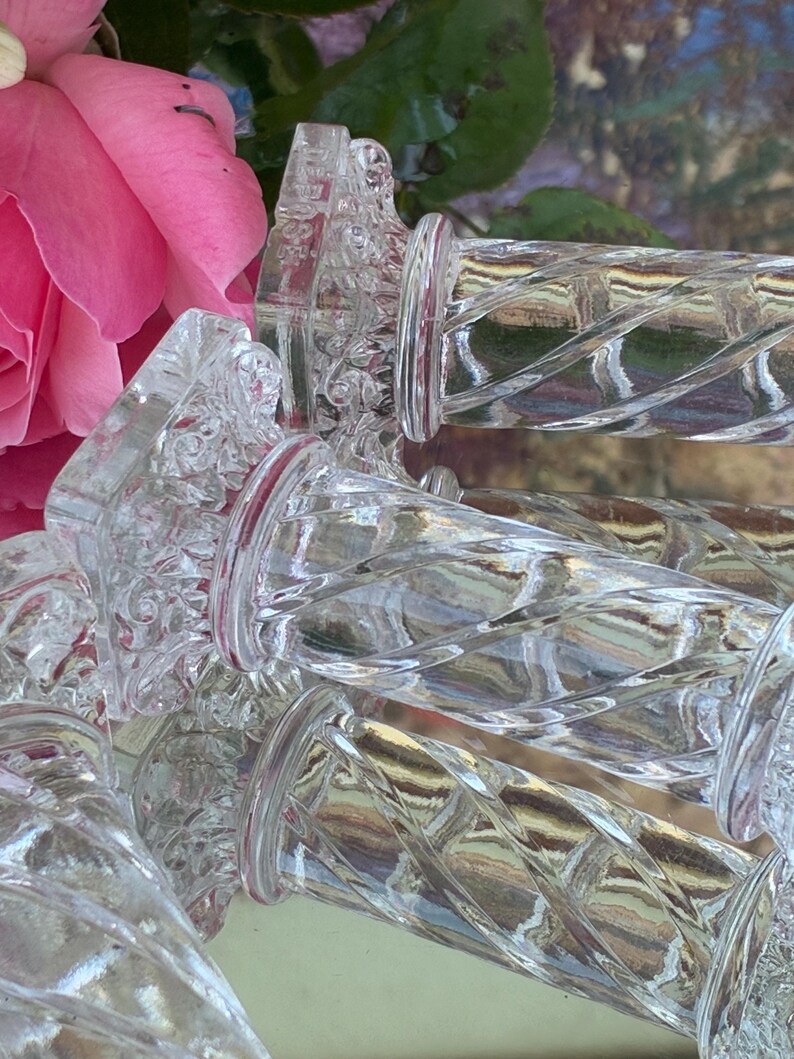 May include: Clear glass bottle stoppers with a decorative, twisted design. The stoppers have a clear, textured body and a red band near the top. A pink rose and green leaves are in the background.