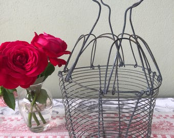Puede incluir: Una cesta de huevos de alambre gris con asa, colocada sobre un mantel a cuadros rojos y blancos. Junto a ella, un pequeño jarrón de cristal contiene rosas rojas brillantes. El fondo es una pared verde claro.