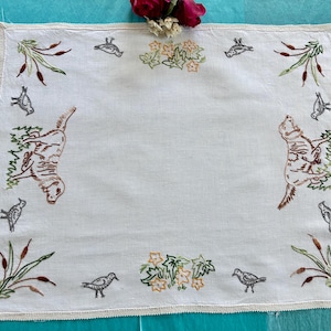 Peut inclure: Une serviette carrée en lin blanc ornée de chiens brodés en marron, de petits oiseaux gris, de massettes vertes et de grappes de fleurs orange. Les bords sont finis par une délicate bordure au crochet blanc, formant un textile décoratif.