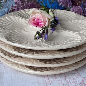 May include: A stack of four off-white ceramic oyster plates. Each plate has a scalloped design with four compartments. A pink rose and purple flowers rest on top of the plates. The plates are on a patterned tablecloth.