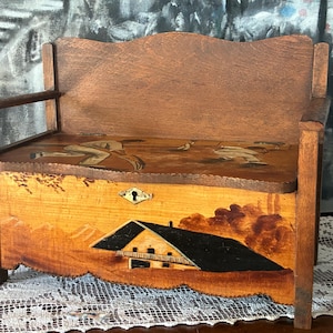 May include: A wooden bench with a painted scene of a house and trees. The bench has a hinged lid and a keyhole. The bench is painted in shades of brown, yellow, and black.