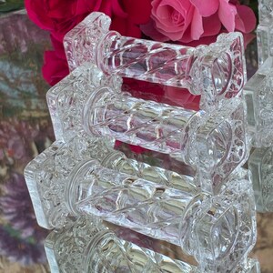 May include: Clear glass decorative objects with a diamond pattern and square ends. The objects are arranged in a stack, reflecting light. The background features pink and red roses, suggesting a vintage or elegant theme.