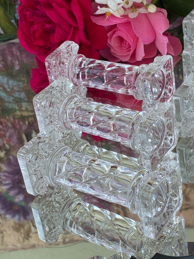 May include: Clear glass decorative objects with a diamond pattern and square ends. The objects are arranged in a stack, reflecting light. The background features pink and red roses, suggesting a vintage or elegant theme.