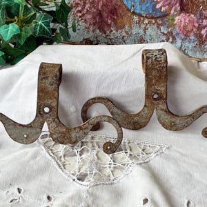 Peut inclure: Deux crochets en métal rouillé d'aspect ancien. Chaque crochet présente une section verticale avec un trou de fixation et une extrémité incurvée en forme de volute. Ils sont posés sur un tissu blanc texturé, évoquant un style vintage ou rustique.
