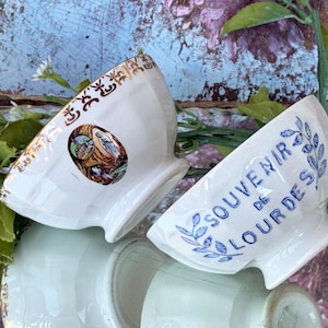Può includere: Due ciotole in ceramica bianca con dettagli decorativi. Una ciotola ha bordi dorati e un'immagine religiosa. L'altra ciotola ha il testo blu "SOUVENIR DE LOURDES". Le ciotole sono inclinate, mostrando l'interno.