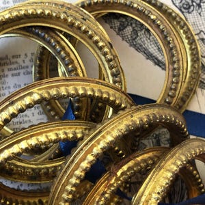 May include: A collection of antique, gold-colored, circular frames with ornate beaded detailing. The frames are arranged in a close-up shot, with a blue ribbon visible. The background includes vintage paper with French text and illustrations.