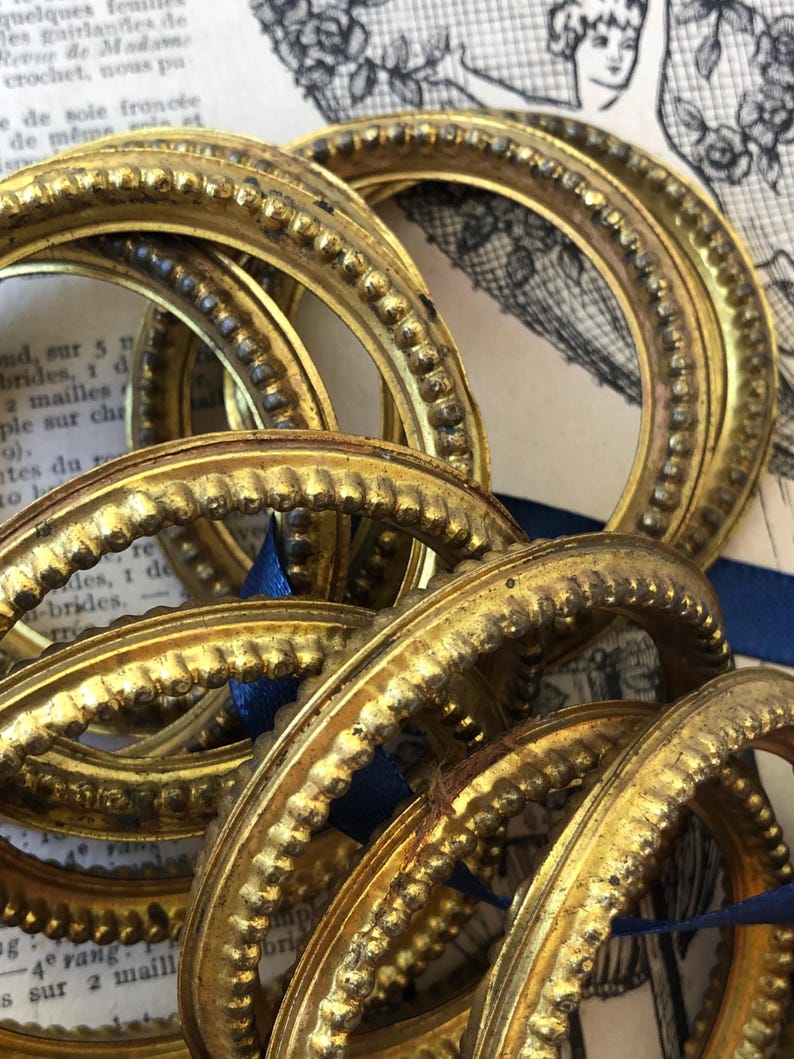 May include: A collection of antique, gold-colored, circular frames with ornate beaded detailing. The frames are arranged in a close-up shot, with a blue ribbon visible. The background includes vintage paper with French text and illustrations.