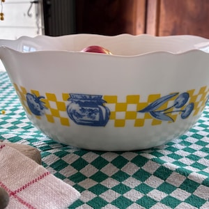 Pode incluir: Tigela de servir de cerâmica branca com borda ondulada. A tigela apresenta uma faixa xadrez amarela e branca com ilustrações azuis de frutas e potes. Está sobre uma toalha de mesa xadrez verde e branca.