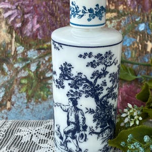 May include: A white porcelain bottle with a blue and white floral design. The bottle has a blue and white floral design on the top and a blue and white scene of a man and a dog in a forest on the front. The bottle is labeled "Lubin" at the bottom.