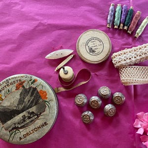 Puede incluir: Una colección de artículos de costura que incluye una lata metálica redonda con la etiqueta Fabrique de Berlingots de Cauterets A. Deltonne, una caja redonda, seis carretes de hilo de colores, dos rollos de encaje crema, seis botones metálicos y una cinta métrica.